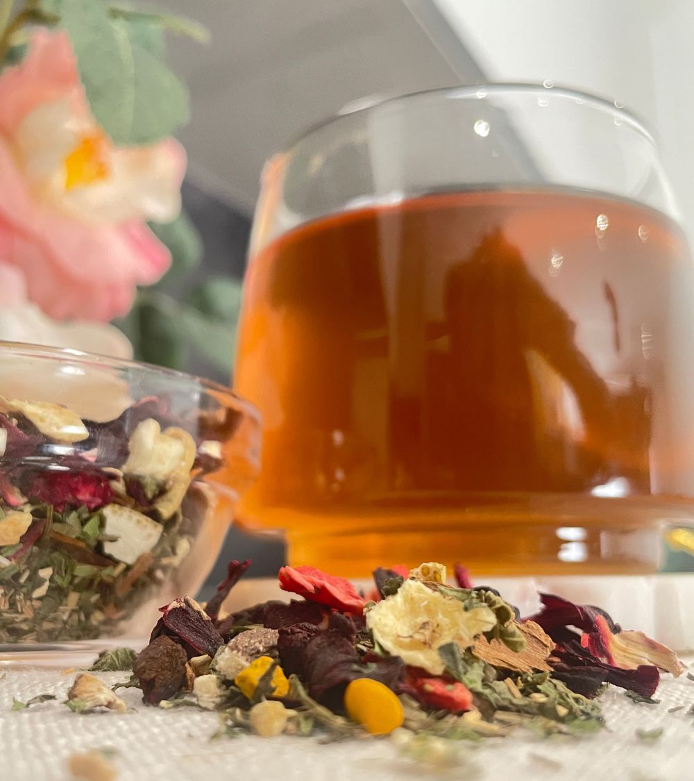A glass cup filled with amber-colored hibiscus zobo bisssap herbal tea, with loose dried flowers and botanicals scattered in front and a small bowl of loose tea leaves on the left.