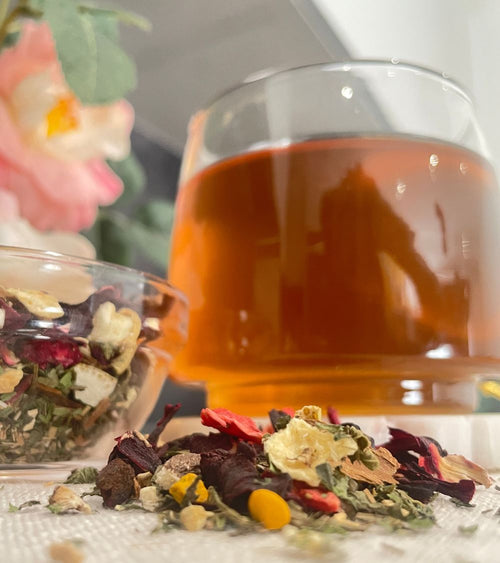 A glass cup filled with amber-colored hibiscus zobo bisssap herbal tea, with loose dried flowers and botanicals scattered in front and a small bowl of loose tea leaves on the left.